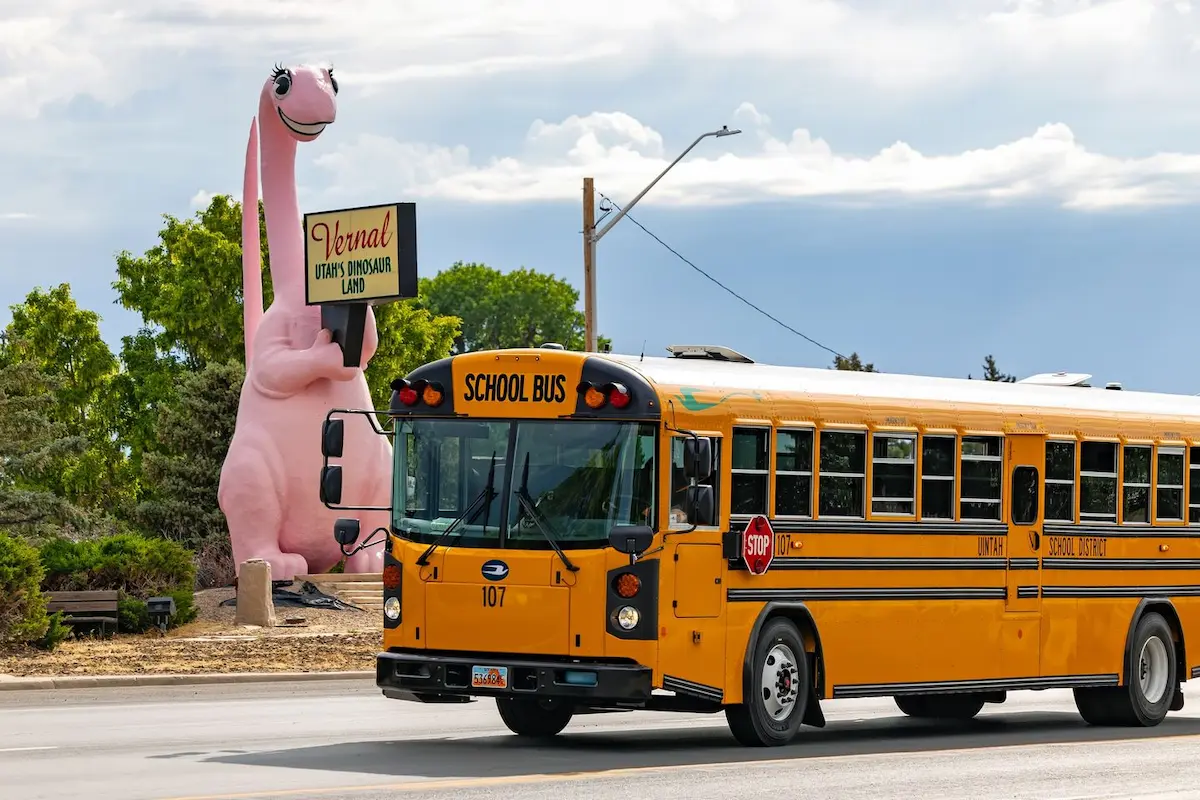 An electric school bus driving in Vernal on a street in front of a big pink dinosaur sign that says "Vernal: Utah's Dinosaur Land"